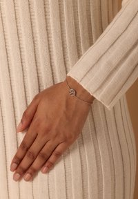 Zilveren armband met een levensboom schittering, gedragen op een hand tegen een textuur van romige, ribbelige stof. Gladde afwerking, delicate ontwerp.