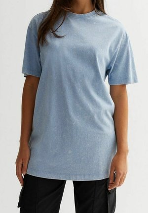 Person wearing a loose, light blue acid-wash short-sleeve shirt with black cargo pants against a plain white background.