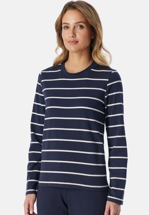Vrouw met schouderlang blond haar, die een marineblauw shirt met lange mouwen en witte horizontale strepen draagt, staat tegen een effen achtergrond.