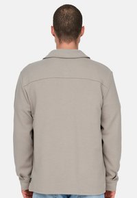 Mann mit kurzen Haaren, der ein hellbeiges langärmeliges Hemd mit Kragen trägt, von hinten vor einem einfarbigen hellen Hintergrund gesehen.