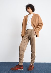 Cárdigan de punto marrón con botones dorados, camiseta blanca, pantalones de chándal beige con franjas laterales y zapatillas color burdeos con acentos de logo.
