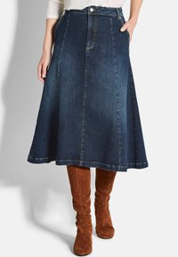 Jupe A-line en jean bleu foncé avec une texture lisse, dotée d'une taille ajustée, de poches avant et d'un ourlet évasé. Portée avec des bottes brunes jusqu'aux genoux.