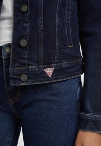Denim jakna u tamnoplavoj boji, sa zlatnim šavovima, zatvaranjem na dugme i trokutastim crvenim "Guess" logom na donjem rubu. Glatka tekstura.