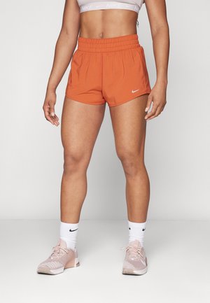 Kobieta ubrana w pomarańczowe sportowe szorty Nike, biały sportowy stanik Nike, białe skarpetki Nike i różowe buty sportowe Nike, stojąca na tle jednolitego tła.