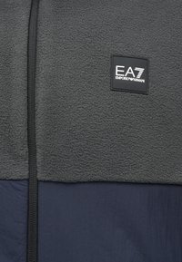 Bunda na zips s tmavošedou fleecovou hornou časťou a námorníckou dolnou časťou. Obsahuje vyšitý štvorec s logom EA7.