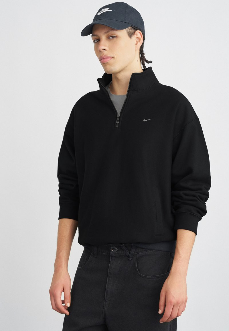Čierna mikina na zips s vysokým golierom a logom Nike, v kombinácii s tmavými džínsami. Mäkká látka a voľný strih, vhodné na voľný čas.