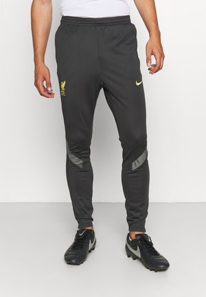 Osoba nosząca czarne spodnie treningowe Liverpool FC z żółtym logo i czarne korki Nike, stojąca na białym tle.