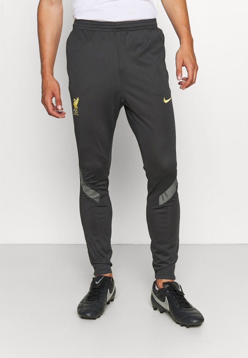 Personne portant un pantalon d'entraînement noir du Liverpool FC avec un logo jaune et des crampons Nike noirs, debout sur un fond blanc.