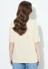 Mujer con cabello castaño largo y ondulado, vestida con una camiseta de manga corta beige y jeans azules, frente a una pared blanca con una sombra proyectada detrás.
