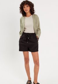 Veste-shirt vert clair, chemise beige, shorts noirs à cordon de serrage et sandales noires. Les shorts ont une taille élastique et des poches latérales.