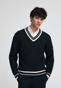 Svart kabelstickad tröja med V-ringning, med vit- och svartrandiga ribbade manschetter och nederkant samt en logotyp på ärmen.