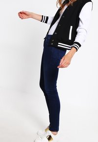 Chaqueta varsity en blanco y negro con puños a rayas, combinada con jeans ajustados azul oscuro y zapatillas blancas con detalles dorados.