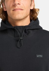 Felpa nera con cappuccio, dotata di un cordoncino e di un'etichetta con logo. Il tessuto sembra morbido, strutturato e aderente attorno al collo.