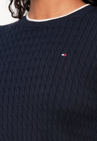 Marineblauer Kabelstrickpullover mit kleinem weißem und rotem rechteckigem Logo auf der Brust, getragen mit weißem Rundhalsa-Shirt.