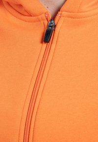 Orange zip-up hættejakke med en sort lynlås, tekstureret stof og synlige stikninger langs kanterne. Minimalistisk design med en tætsiddende pasform.