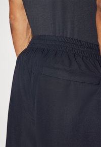 Nahaufnahme der Taille einer Person, die schwarze elastische Hosen mit einem Rückentasche und einem eingesteckten schwarzen Hemd trägt.