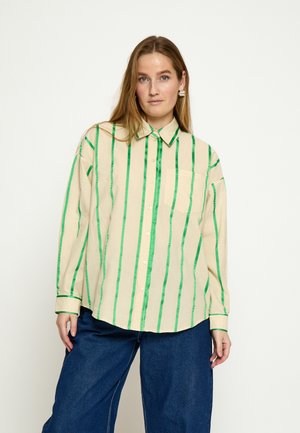 Žena oblečená v béžovej blúzke s dlhými rukávmi a vertikálnymi zelenými pruhmi a tmavomodrých širokých nohaviciach stojaca pred jednoduchým svetlým pozadím.