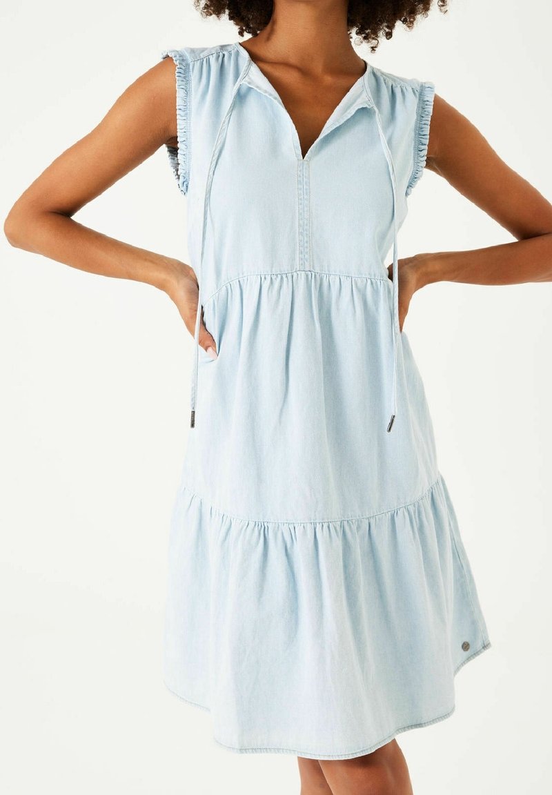 Jasnoniebieska sukienka jeansowa z warstwowym fasonem, krótkimi rękawami i dekoltem w serek. Sznurowany detal przy szyi oraz falbanki na rękawach.