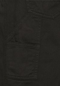 Detailní záběr na černou denkovou látku s viditelným prošíváním a švy kapes s ostrými designovými prvky.