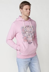 Roza kapuca s grafičnim dizajnom lobanje, ki vključuje tropske elemente. Ima kenguru žep in vrvice na kapuci, izdelana je iz mehkega materiala.