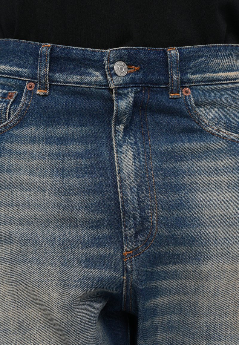 Närbild av blårandiga denimjeans vid midjan med knapp, bältesöglor och framfickor på svart bakgrund.
