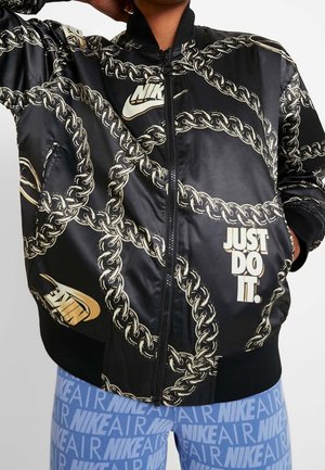 Veste zippée noire Nike avec motif chaîne dorée, logos Nike, et texte "Just Do It.", portée avec des leggings Nike Air bleus.