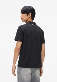 Čierne polo tričko s golierom, texturovaná tkanina a logo na chrbte. V kombinácii so svetlosivými, mierne opranými džínsami.