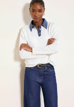 Woman with arms crossed wearing a white sweater with a denim collar, blue jeans, and a black belt with a gold buckle.