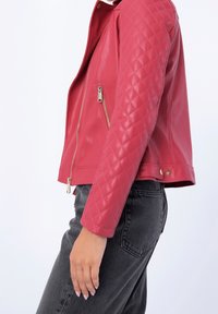 Červená faux kožená bunda s prošívanými rukávy, postranními kapsami na zip a zlatým kováním. Úzký střih s hladkou texturou.