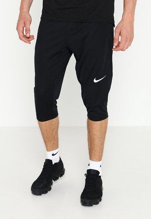 Man draagt zwarte Nike cropped broek, zwarte Nike schoenen en witte sokken met zwarte Nike-logo's, staand op een effen witte ondergrond.