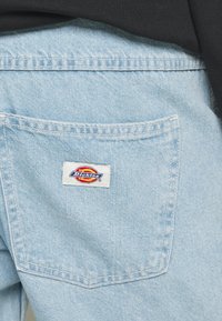 Vaaleansiniset farkut, joissa on takatasku, jossa on suorakulmion muotoinen etiketti, jossa on "Dickies"-logo punaisena, keltaisena ja sinisenä.