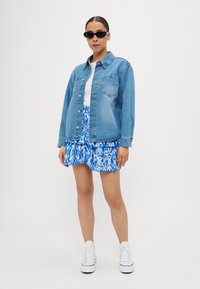 Denim jakna v svetlo modri barvi z žepom na prsih, oblečena preko bele majice in modre patterned krilce z volančki, v kombinaciji z belimi supergami.