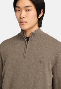 Maglione marrone con colletto a zip, texture a coste e logo ricamato sul petto. Tessuto morbido e design aderente.