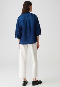 Giubbotto in denim blu scuro con vestibilità ampia, polsini a due bottoni e dettaglio a yoke. Abbinato a pantaloni bianchi a gamba dritta e scarpe nere.