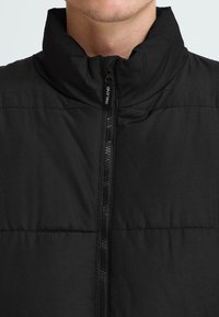Sort pufferjakke med høj krave, frontlukning med lynlås og quiltet tekstur. Stoffet virker glat og vandafvisende. Minimalistiske detaljer.