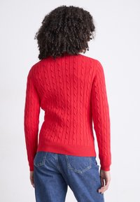 Personne aux cheveux bouclés portant un pull rouge en maille torsadée et un jean bleu, debout de dos face à l'appareil photo sur un fond uni.