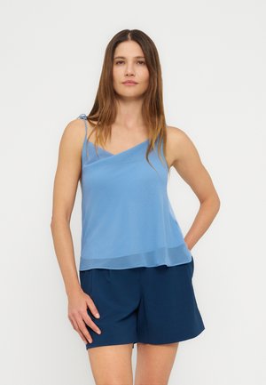 Donna con top asimmetrico a spalline azzurro chiaro e pantaloncini blu scuro, in piedi con una mano in tasca contro uno sfondo semplice.
