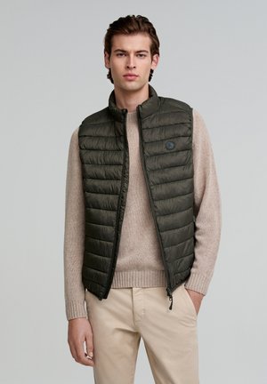 Donkergroene gewatteerde vest met een rits aan de voorkant en hoge kraag, gedragen over een lichtbeige trui. Bevat een ronde logo op de linkerborst.