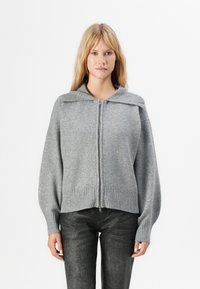 Blond kvinne iført en grå zip-up genser med bred krage og teksturerte mørke bukser, som står mot en enkel hvit bakgrunn.