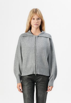 Femme blonde portant un pull zippé gris avec un large col et un pantalon texturé foncé, se tenant devant un fond blanc uni.