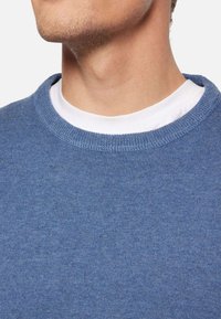 Blauer Rundhals-Pullover aus weichem Strickmaterial, mit einem gerippten Halsausschnitt, über einem weißen T-Shirt getragen.