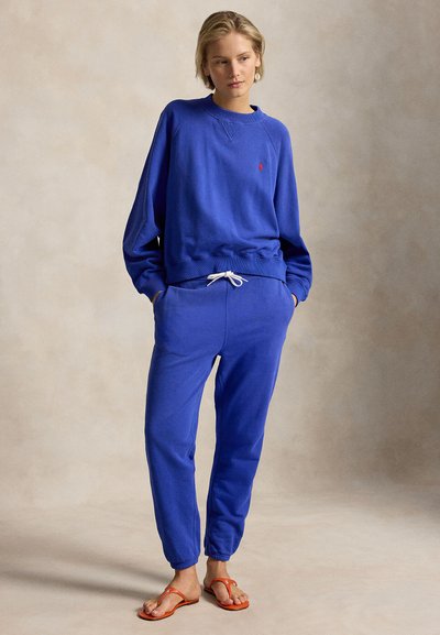 Γυναίκα που φοράει μπλε φούτερ και αντίστοιχη φόρμα με τα χέρια στις τσέπες, όρθια ξυπόλυτη σε ουδέτερο φόντο.