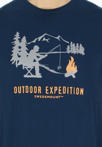 Marineblå t-shirt med en grå fiskescene, der har træer og et bål. Trykt tekst med ordene "UDENDØRS EKSPEDITION" i orange. Blødt materiale.