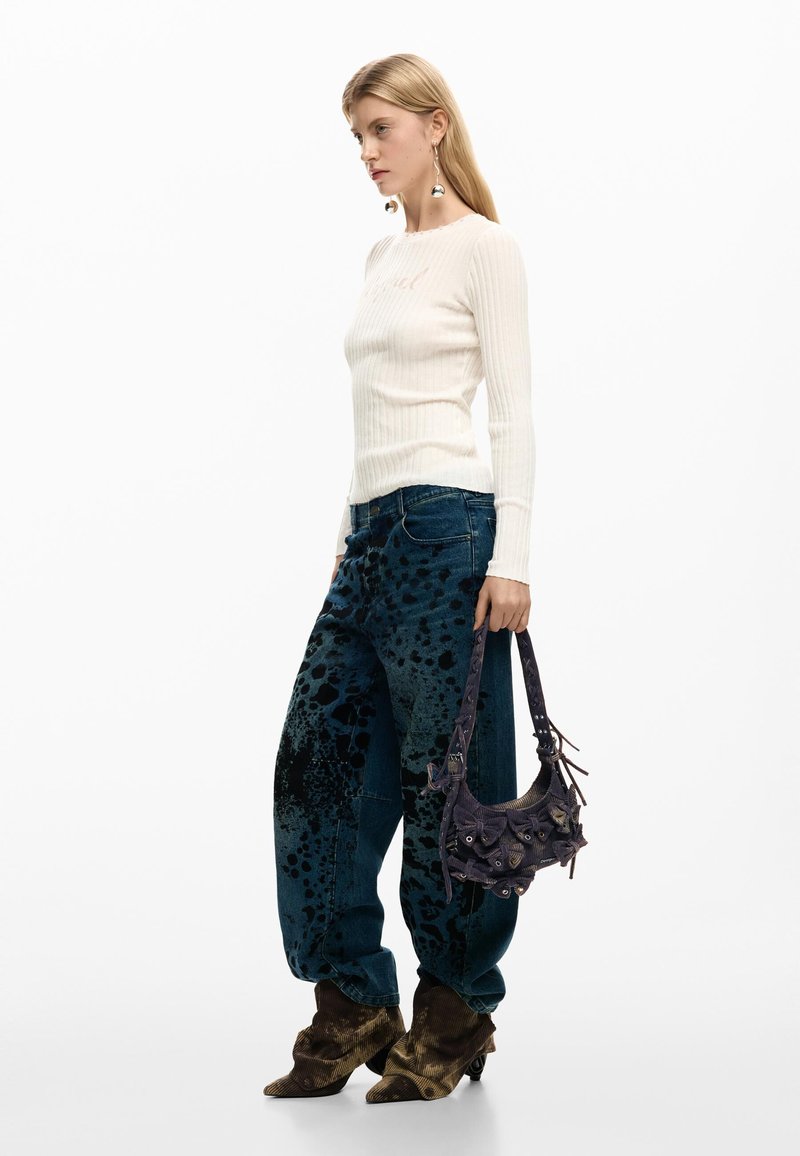 Mujer con cabello largo y rubio, vestida con un top blanco acanalado de manga larga, vaqueros holgados con estampado de salpicaduras azules, botas marrones, sosteniendo un bolso de mano morado oscuro con detalles florales.