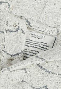 Vitt frottétyg med en mjuk textur, med ett subtilt grått mönster. Etiketten visar varumärket "EMMAMALENA" och skötselråd.
