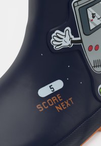 Detalle de un zapato azul marino oscuro con un gráfico de un robot de dibujos animados y texto en naranja que muestra "SCORE NEXT" y el número 5 en un óvalo blanco.