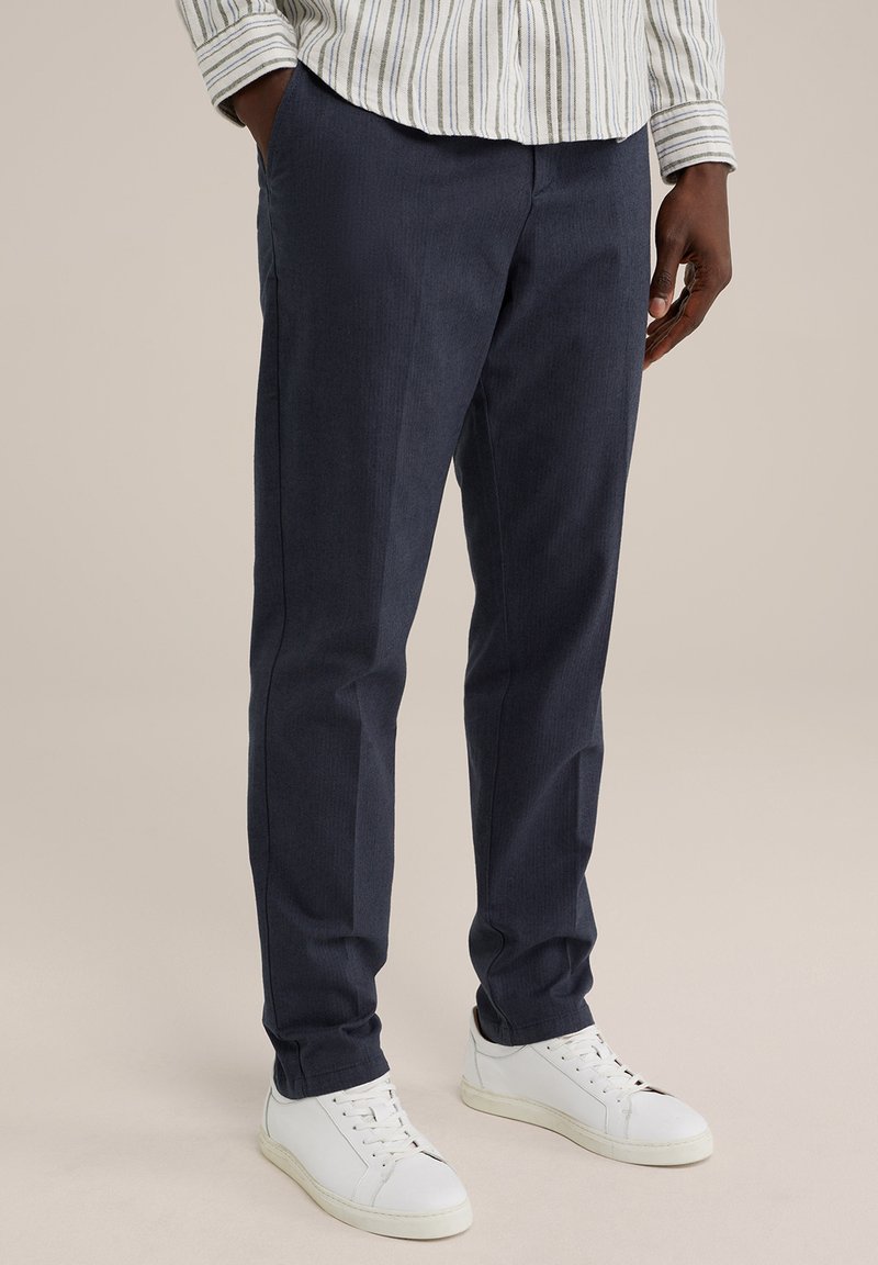 Dark navy trousers in a textured fabric with a straight cut, paired with white sneakers. The waistband is flat, and pockets are included.