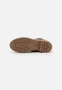Botte en suède marron avec une semelle en caoutchouc présentant un motif de bande de roulement texturé, un détail de sangle ajustable et une empreinte de logo sur la semelle.