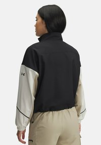 Femme aux cheveux noirs portant une veste color-block noire et beige avec des poignets élastiques et un pantalon beige, faisant face à l'arrière de l'appareil photo.