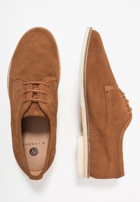 Chaussures habillées en daim marron avec un design à lacets, dotées d'une semelle lisse et d'accents de couture subtils. L'intérieur présente un logo de la marque.